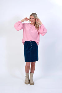 Woman in pink balloon sleeve top, blue button-front skirt, and beige ankle boots on white background