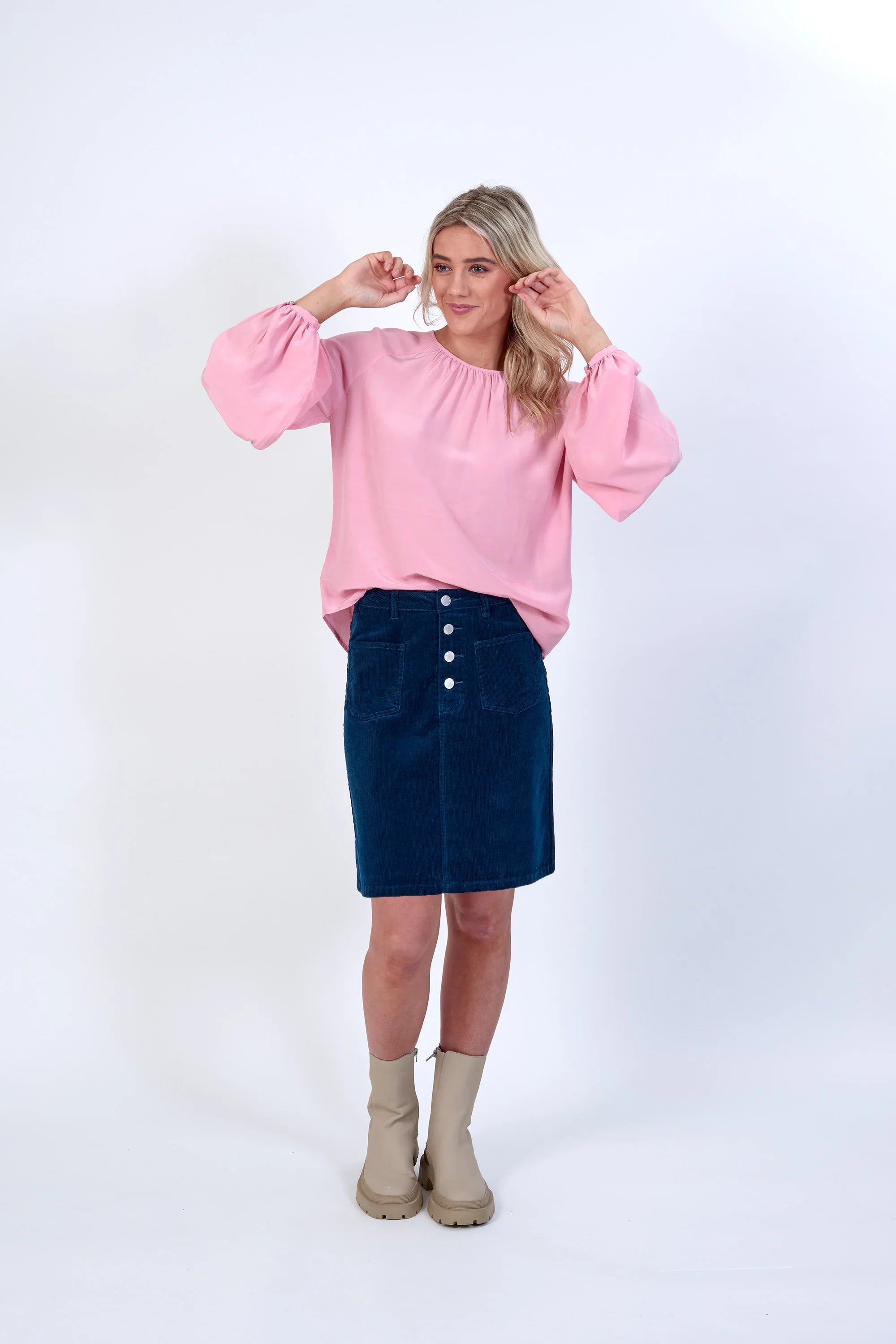 Woman in pink balloon sleeve top, blue button-front skirt, and beige ankle boots on white background