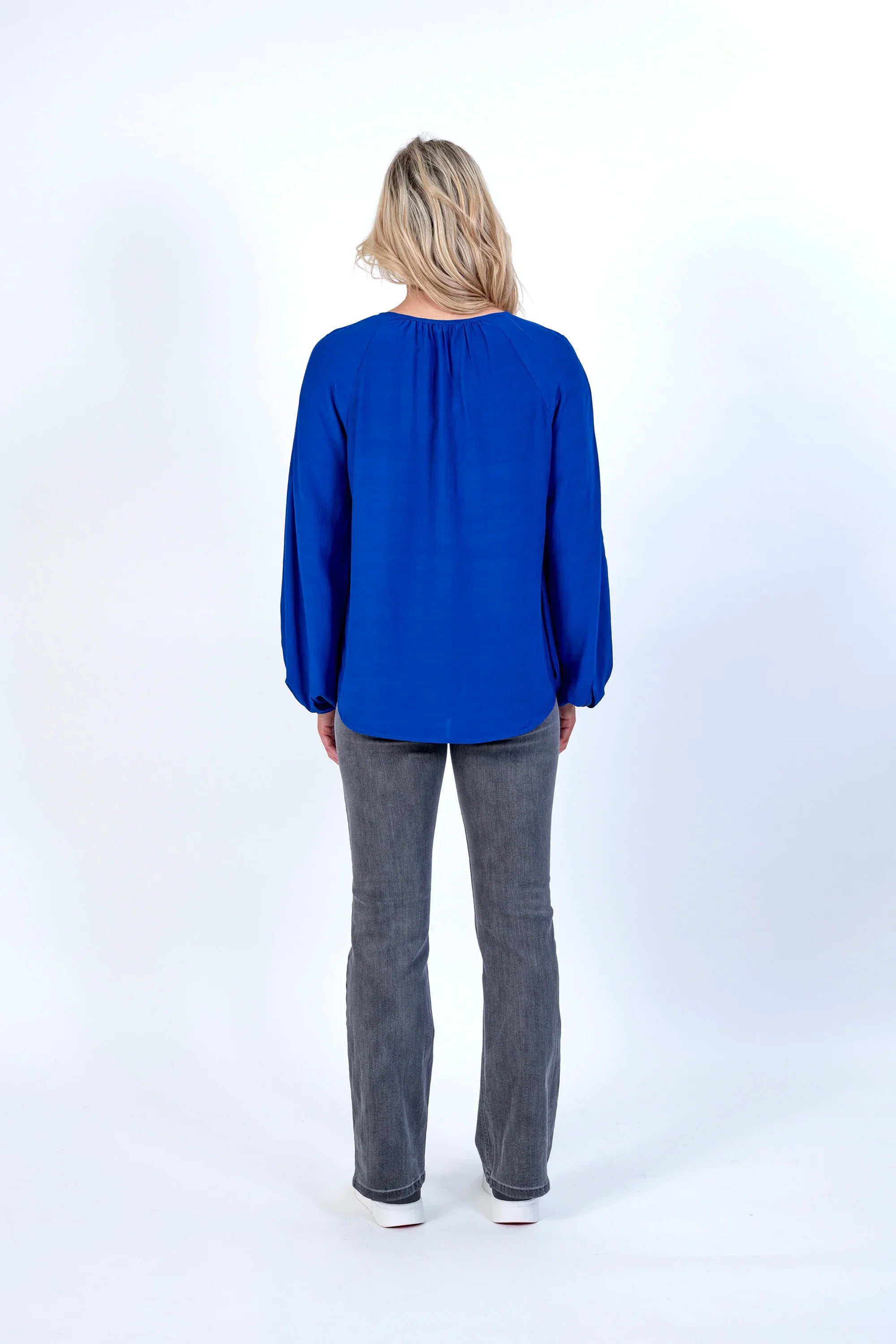Woman wearing electric blue long sleeve top and grey jeans, back view, studio setting