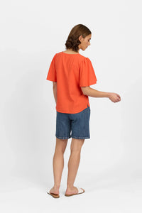 Woman in loose orange short sleeve top and denim shorts, back view, studio setting