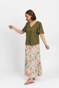 Woman in olive short sleeve V-neck top and long floral skirt with sandals, standing against white background