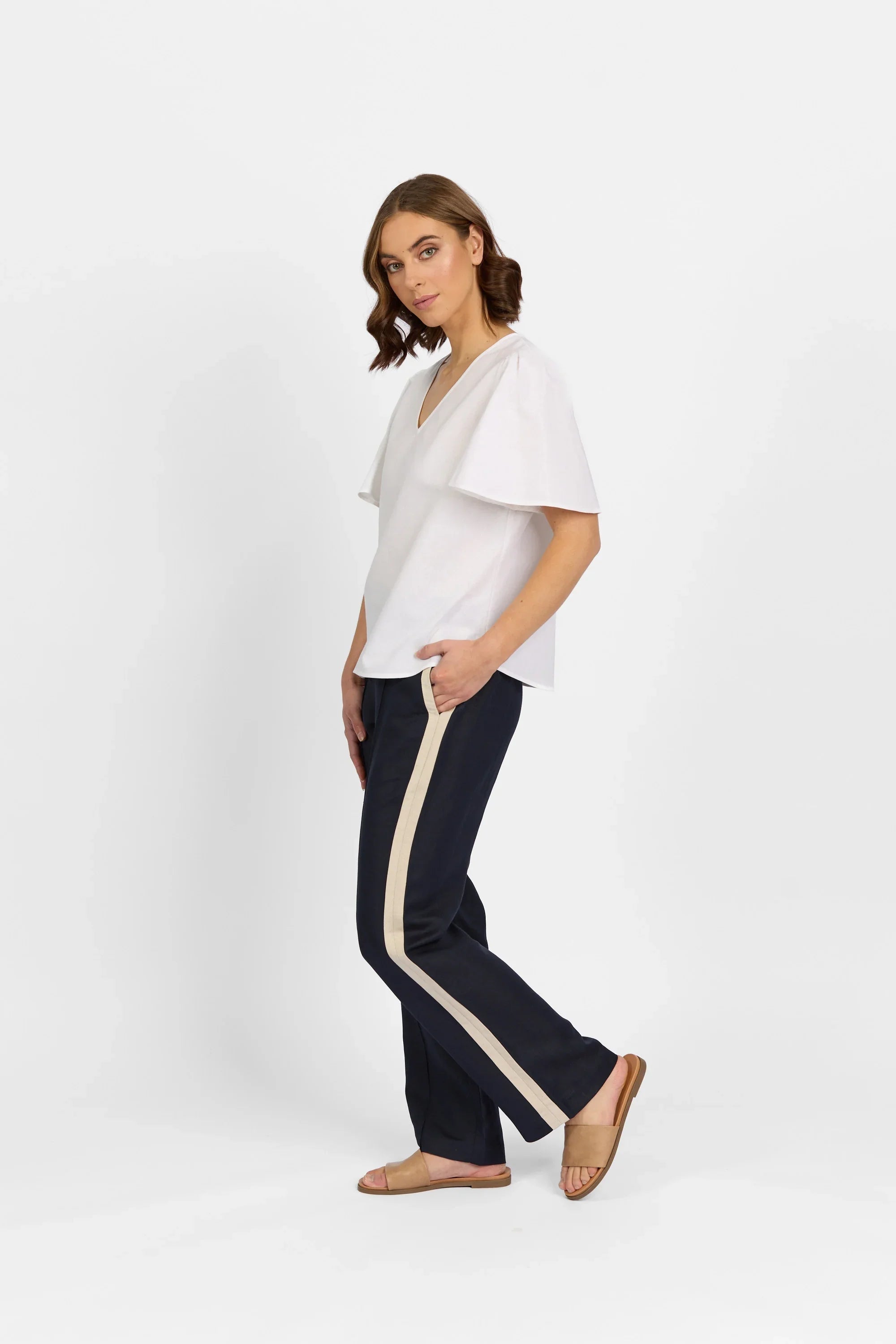 Woman wearing white flutter sleeve top and navy pants with beige stripe on white background
