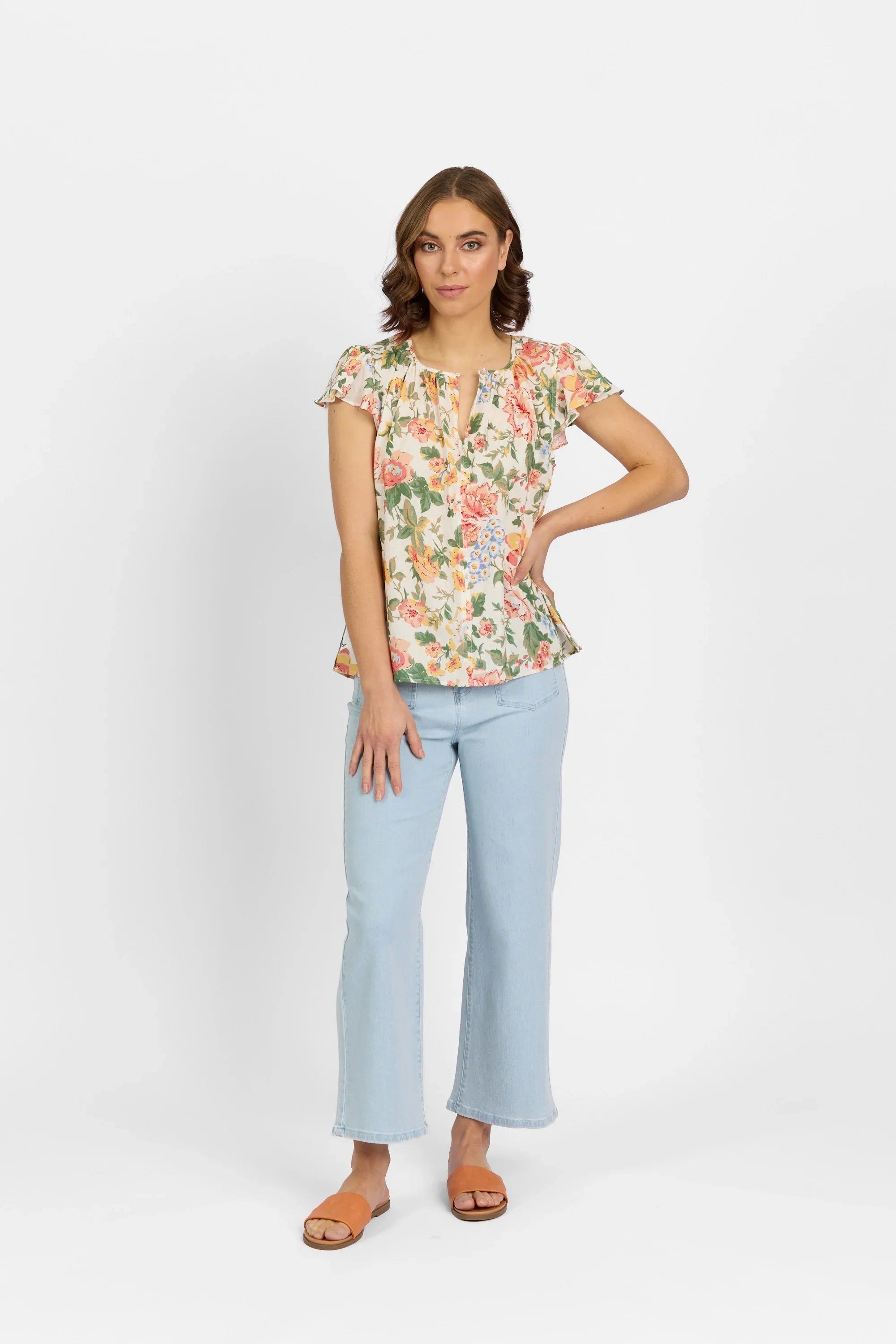 Woman in floral flutter sleeve blouse, light wash jeans, and orange sandals, standing against white background