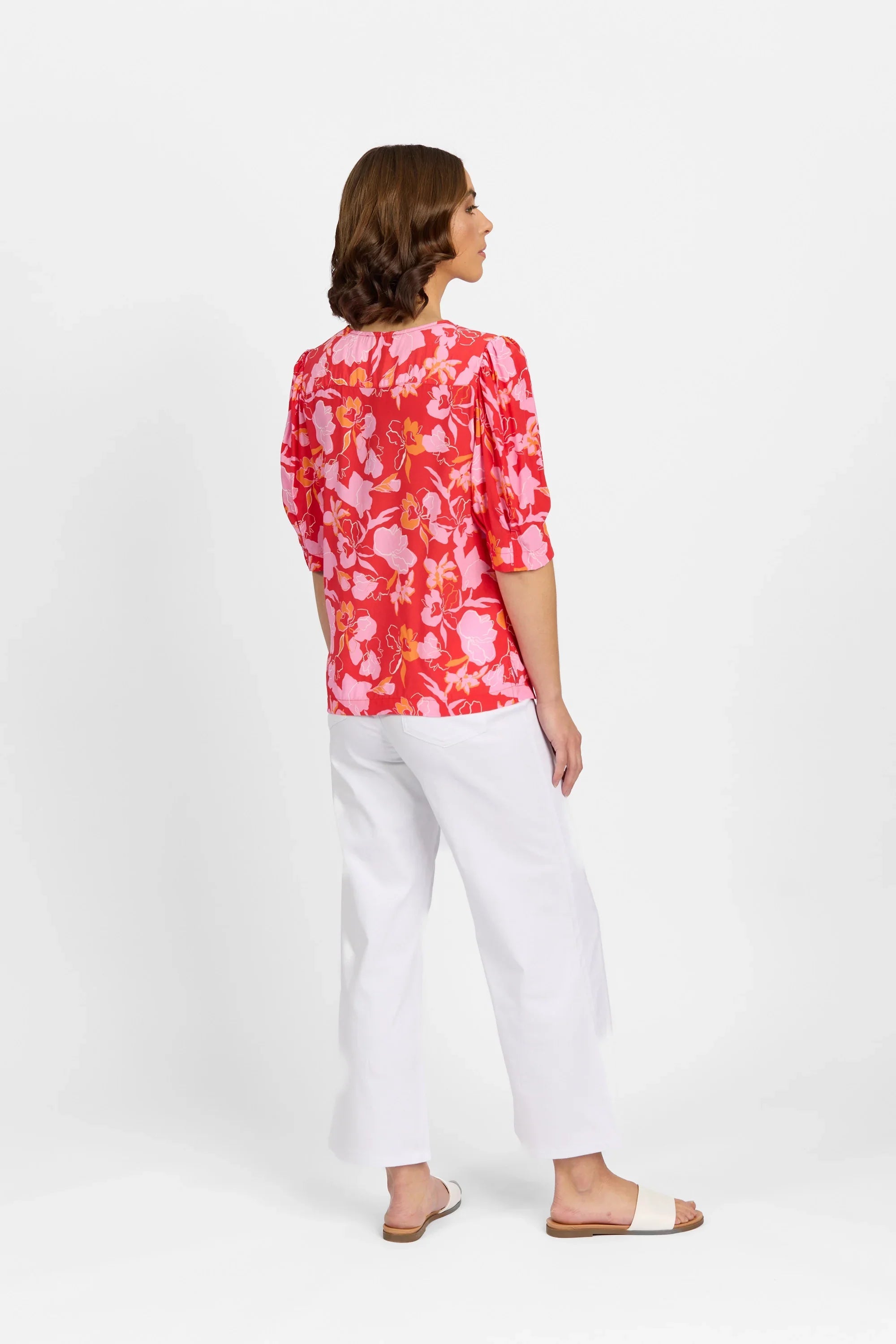 Woman wearing a pink floral top with puff sleeves and white pants, back view, studio shot.