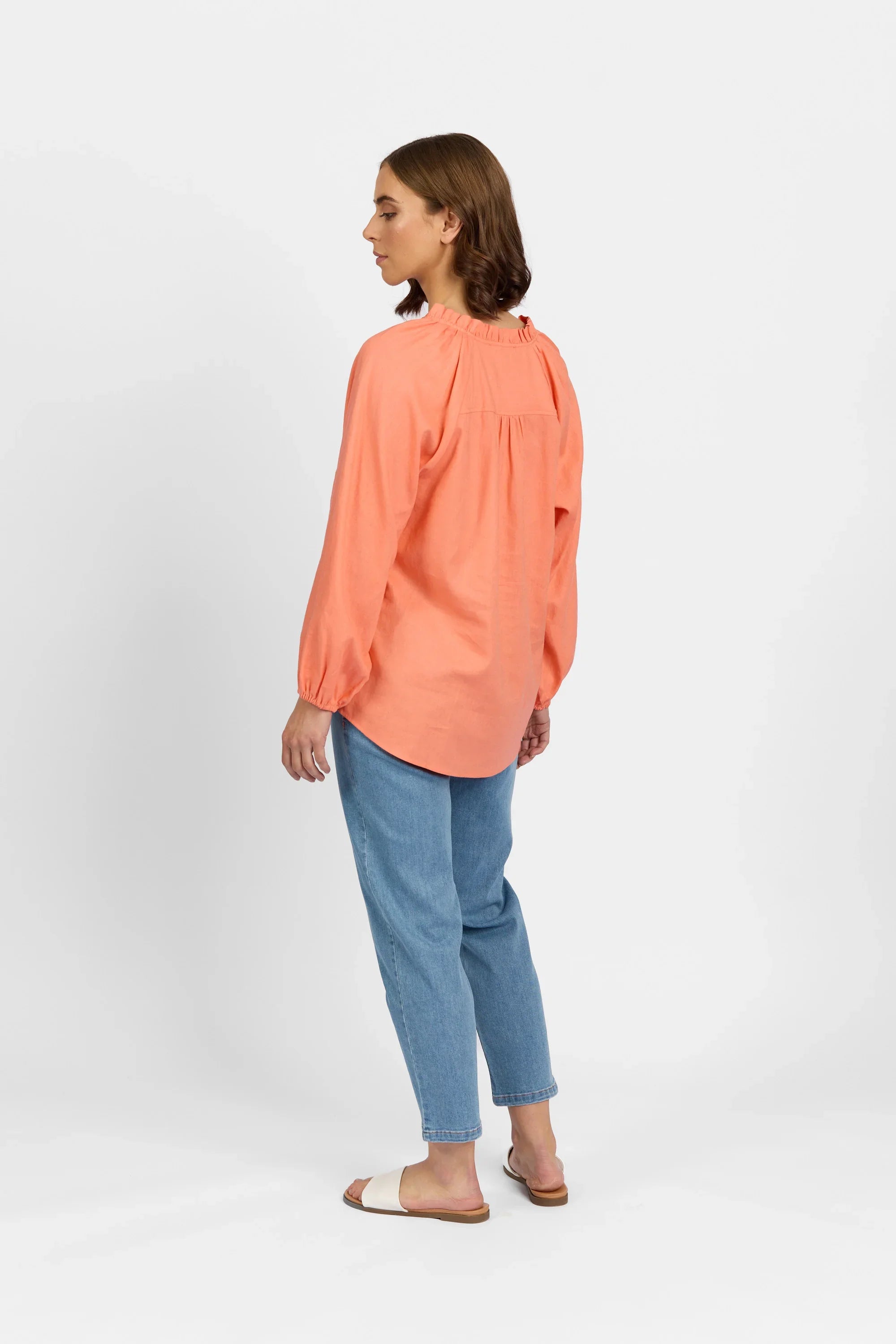 Woman wearing a coral long-sleeve shirt with blue jeans and sandals, standing sideways