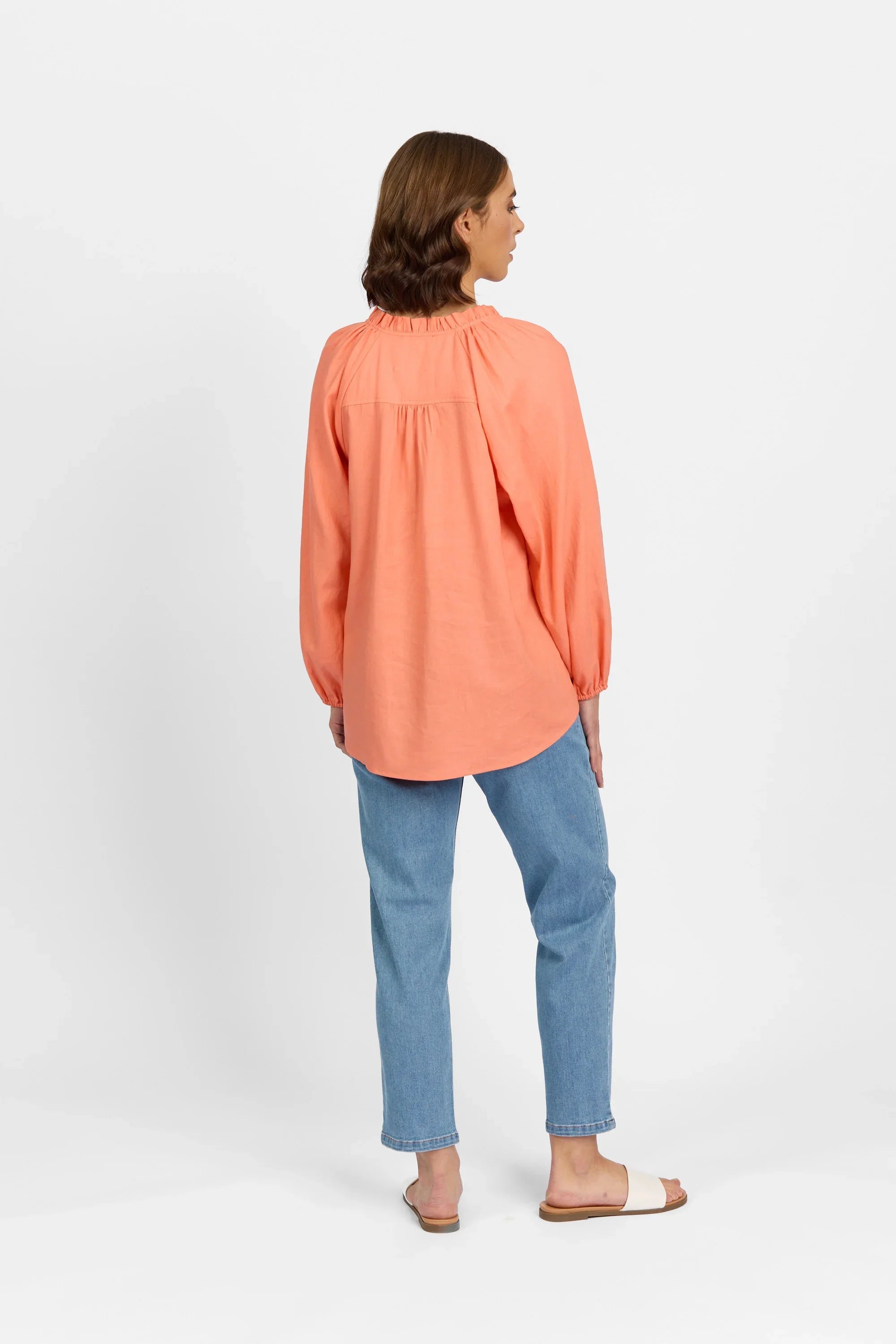 Woman wearing a coral long sleeve blouse, blue jeans, and white sandals, shown from behind.