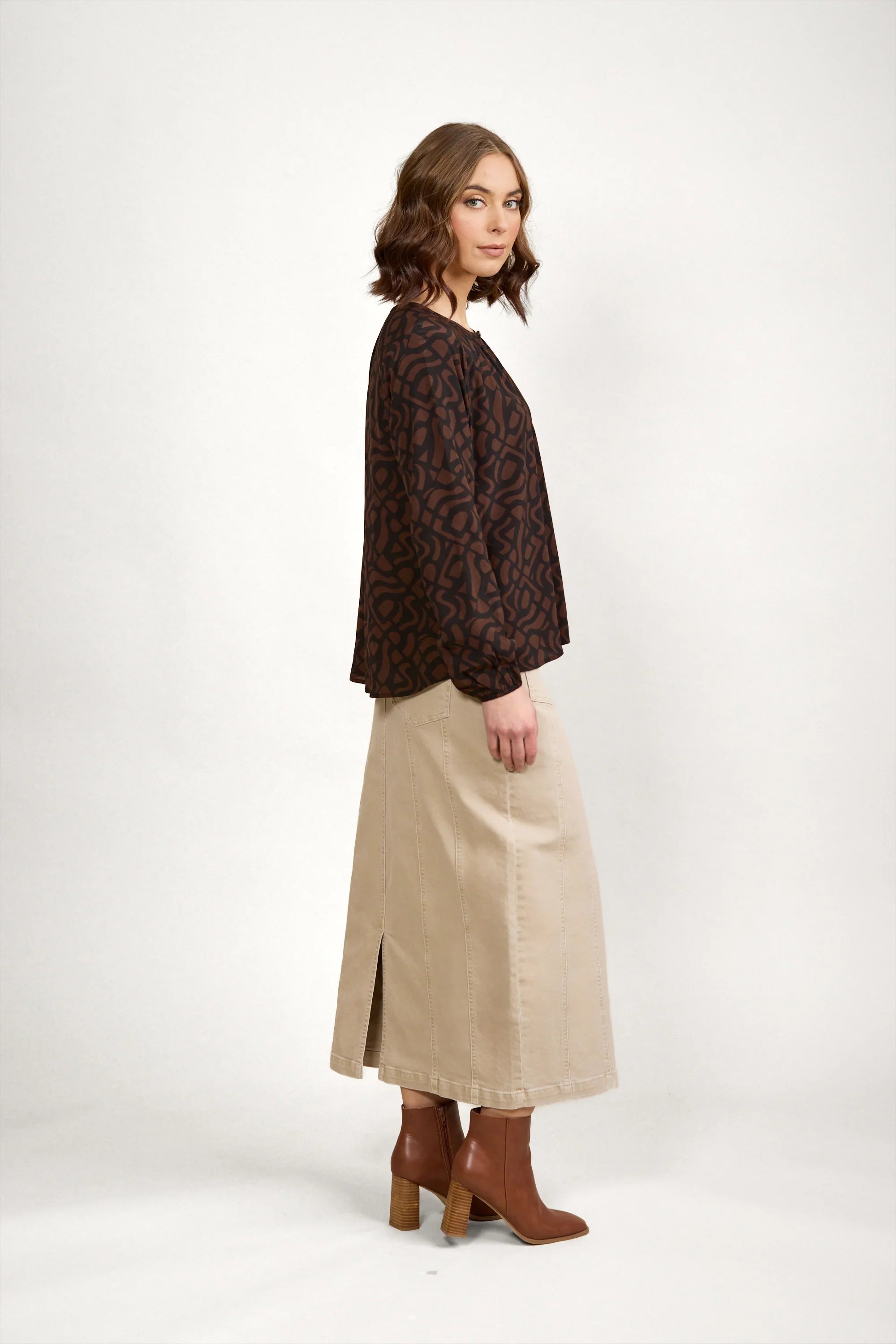 Woman in brown patterned top, beige maxi skirt, and brown ankle boots on white background