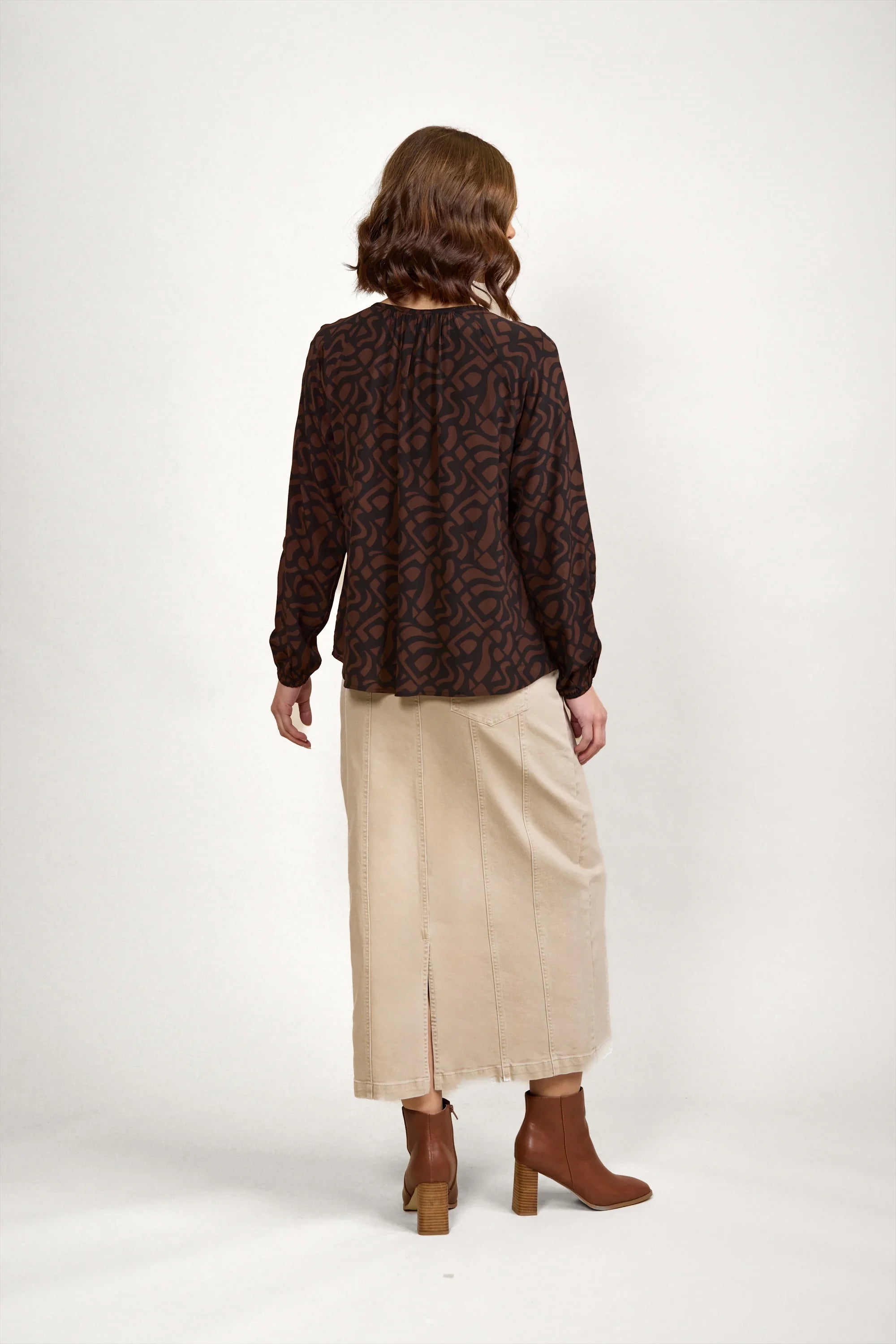 Woman wearing brown patterned long sleeve top, beige midi skirt, and brown heeled boots, back view