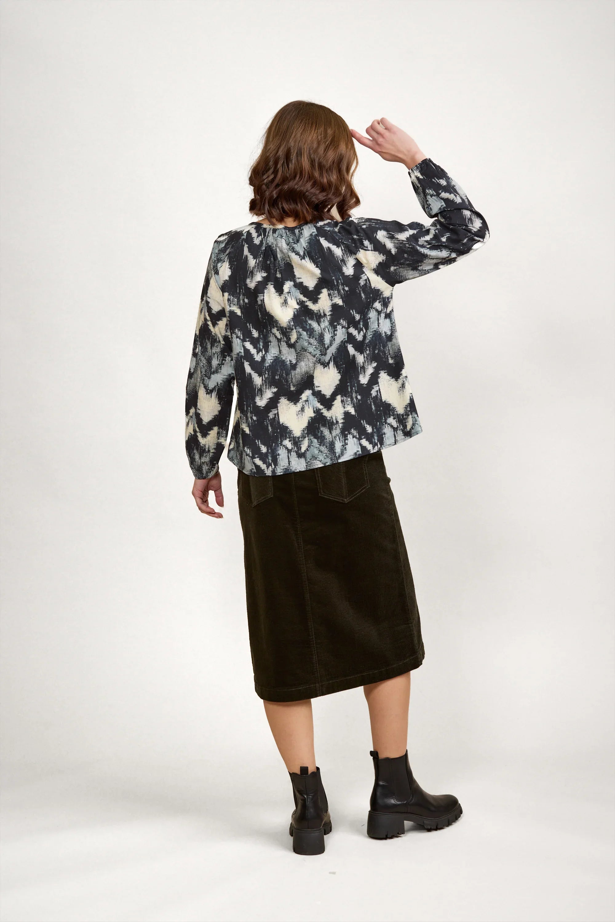 Woman in patterned long sleeve blouse, black midi skirt, and ankle boots, back view