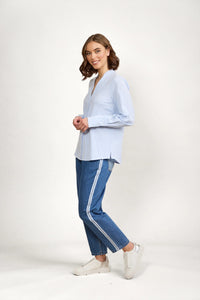Woman wearing a blue and white striped button-up shirt, blue jeans with side stripe, and white sneakers, posing against a plain background.
