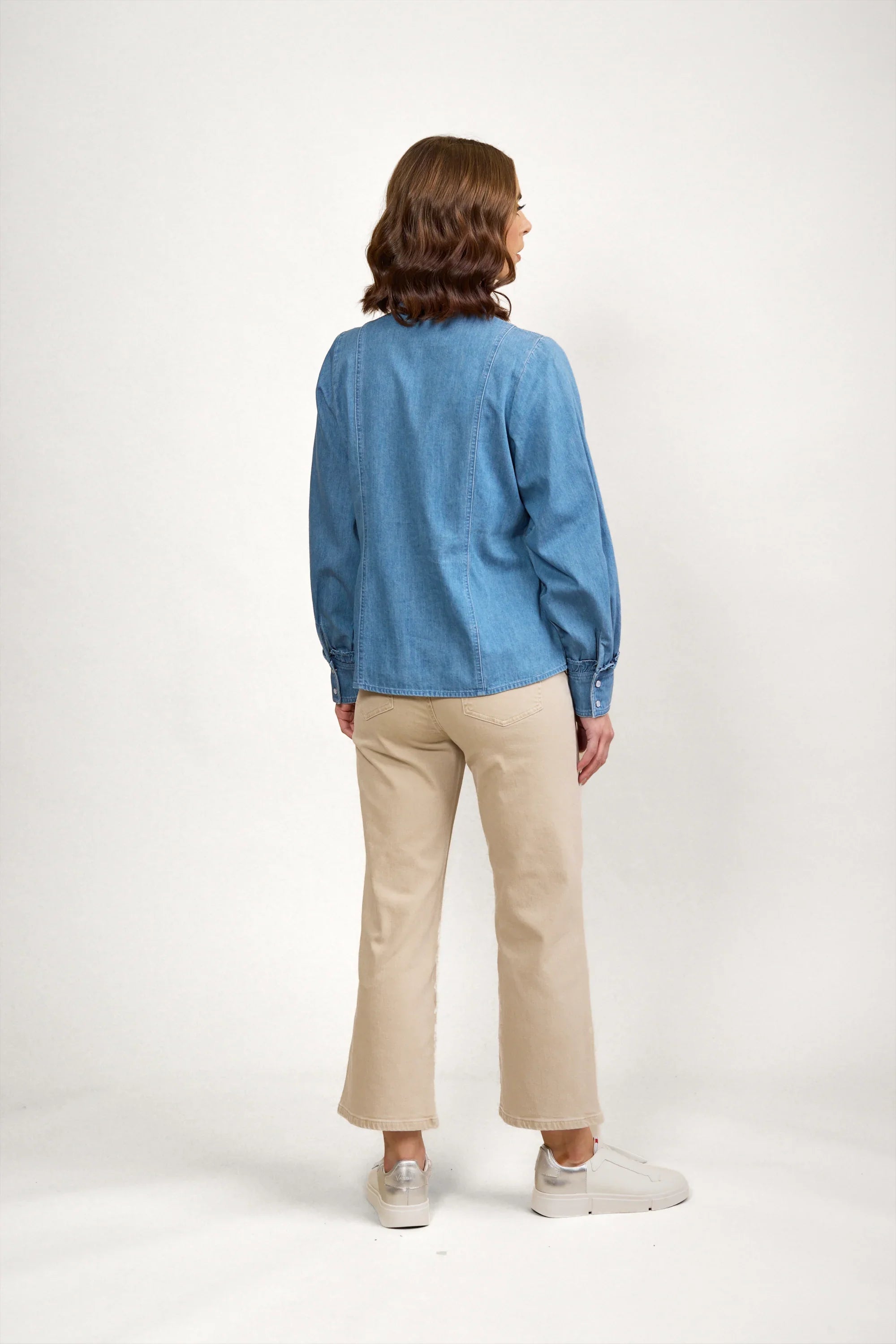 Woman wearing a blue denim shirt and beige pants, shown from the back on a plain background.