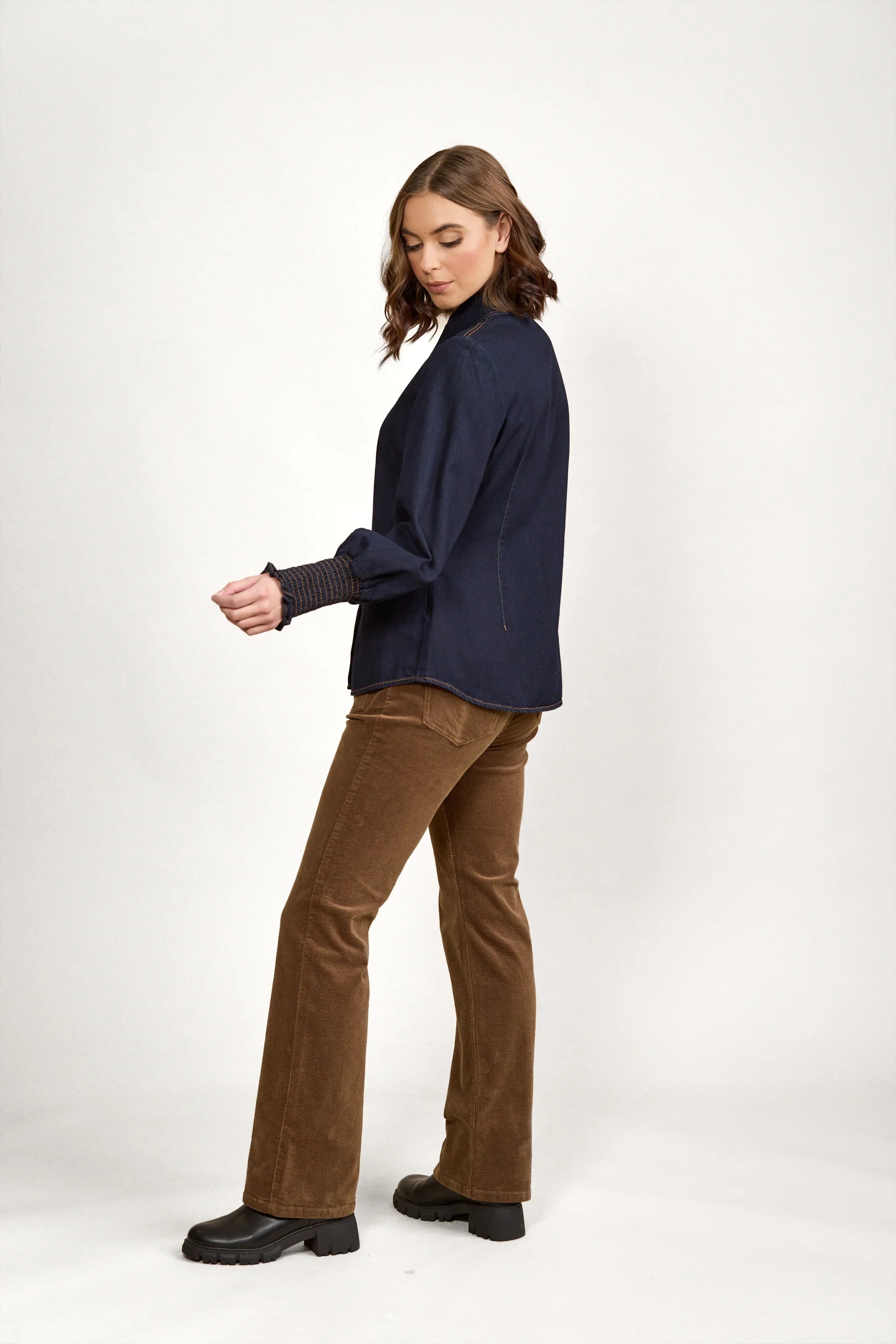Woman wearing dark denim shirt, brown corduroy pants, and black boots on white background