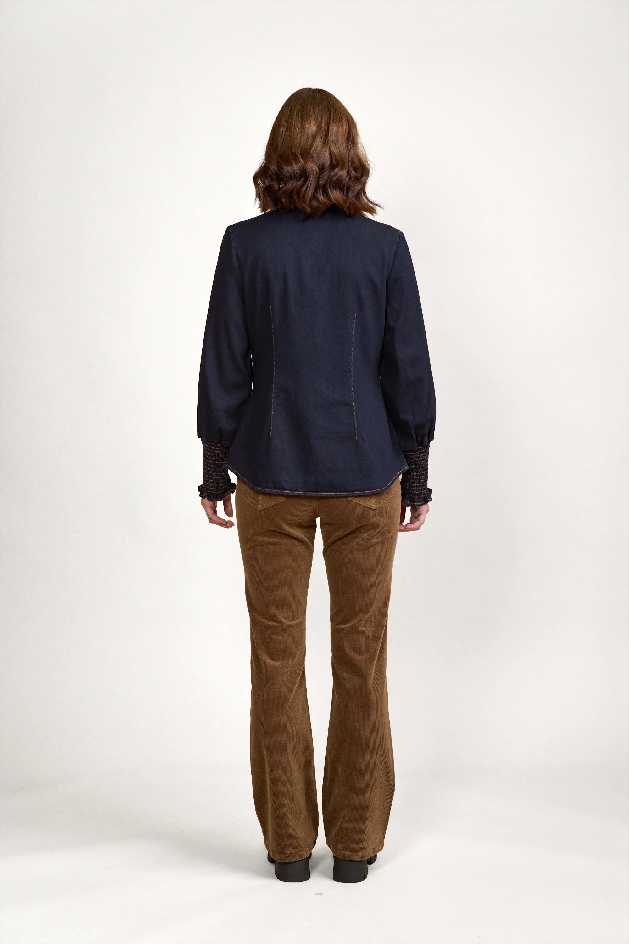 Woman in dark denim shirt with puff sleeves, brown corduroy pants, black boots, rear view