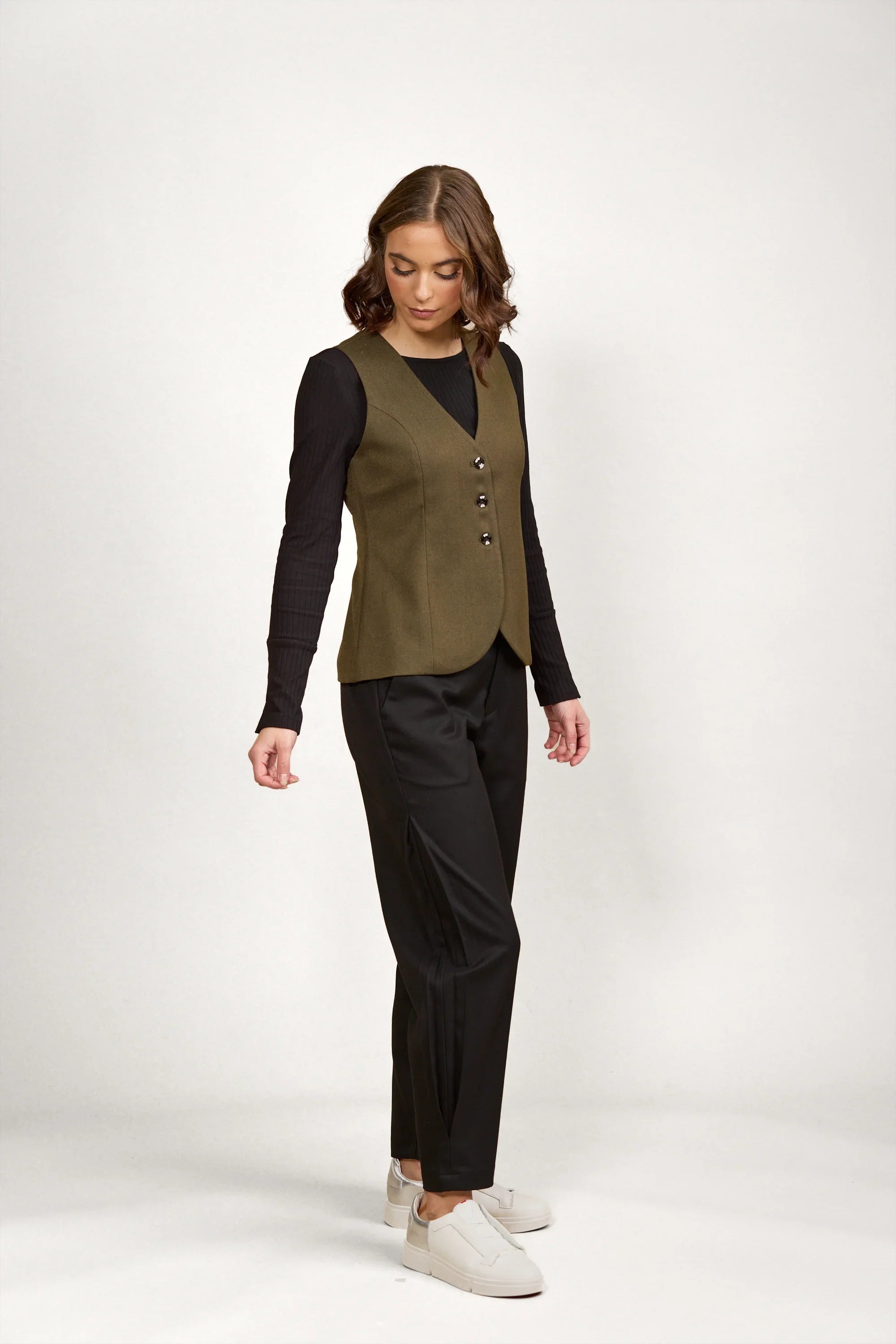 Woman wearing an olive green buttoned vest, black long sleeve top, black pants, and white sneakers