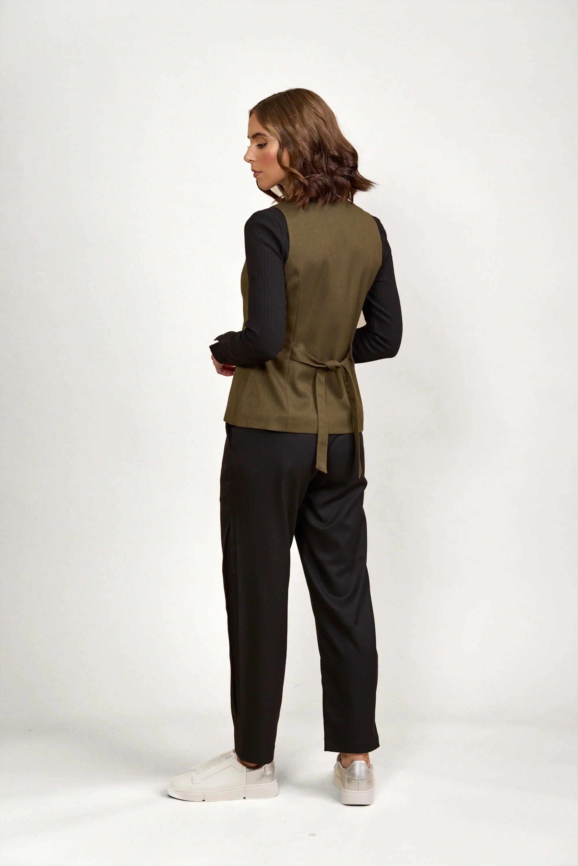 Woman wearing olive green vest with tie back, black trousers, and white sneakers