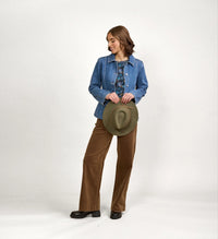 Woman modeling blue denim jacket with brown wide-leg pants, holding a green hat