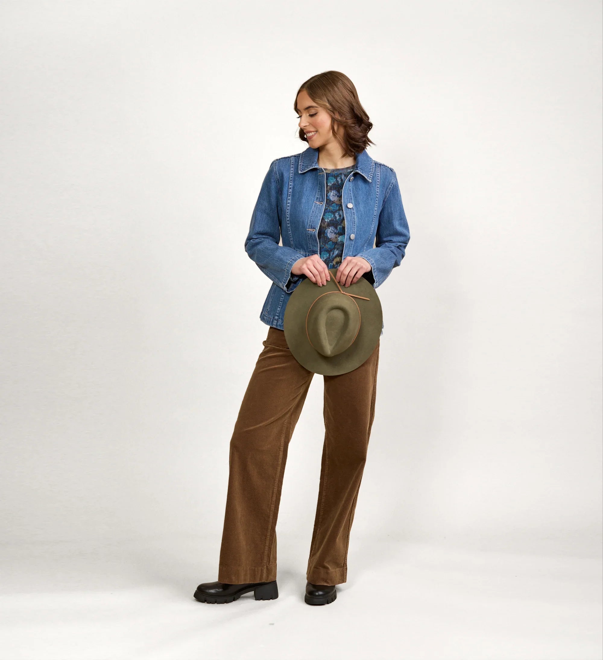 Woman modeling blue denim jacket with brown wide-leg pants, holding a green hat