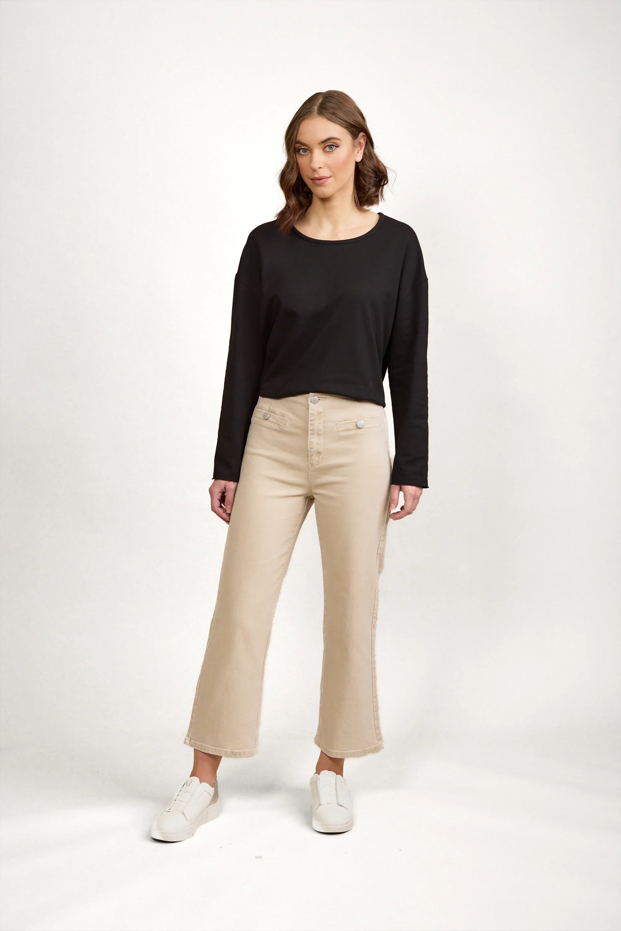 Woman wearing beige wide leg jeans, black long sleeve top, and white sneakers on white background