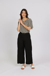 Woman in wide-leg black pants, geometric print blouse, and white slides, studio background