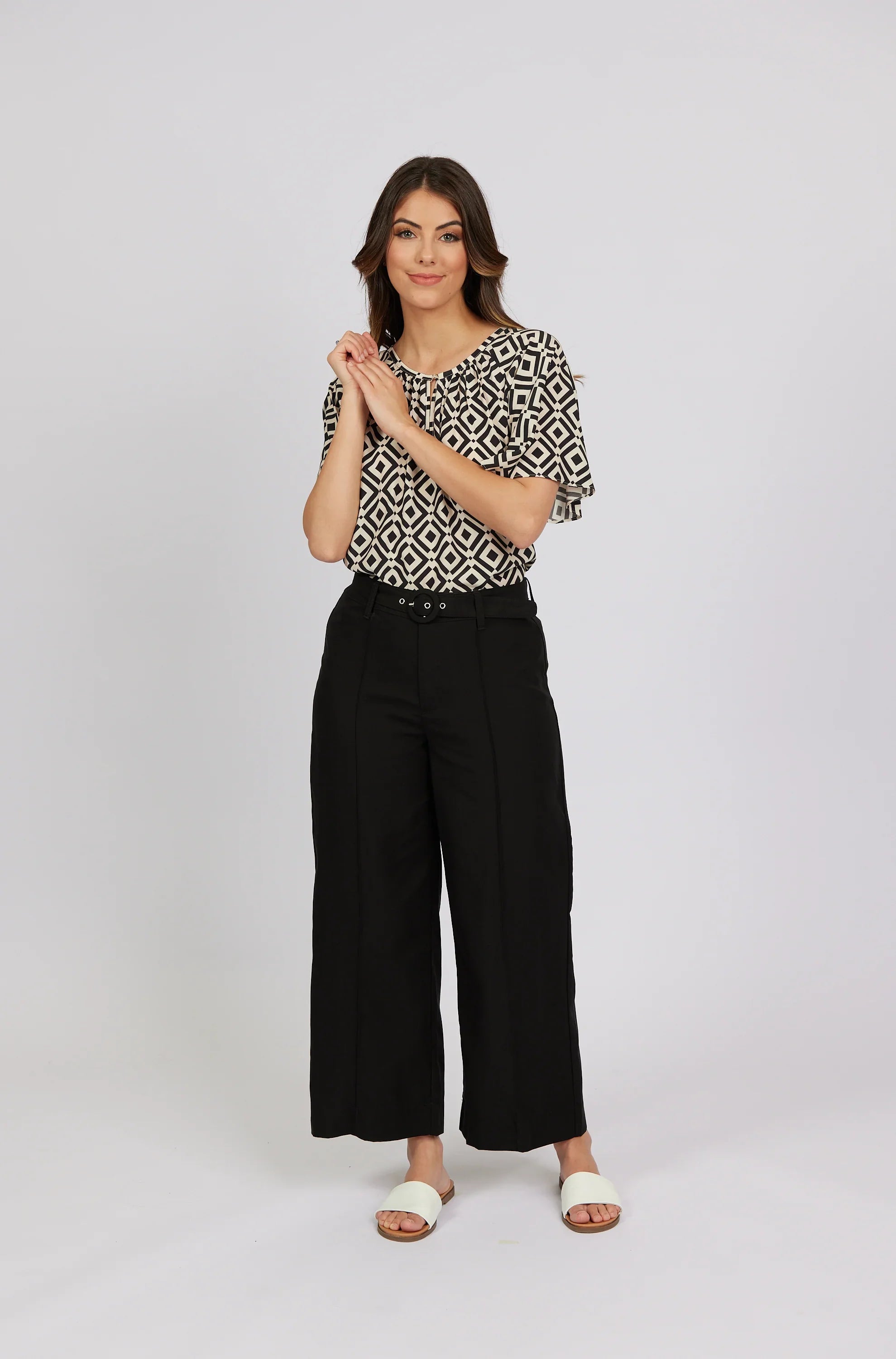 Woman in wide-leg black pants, geometric print blouse, and white slides, studio background