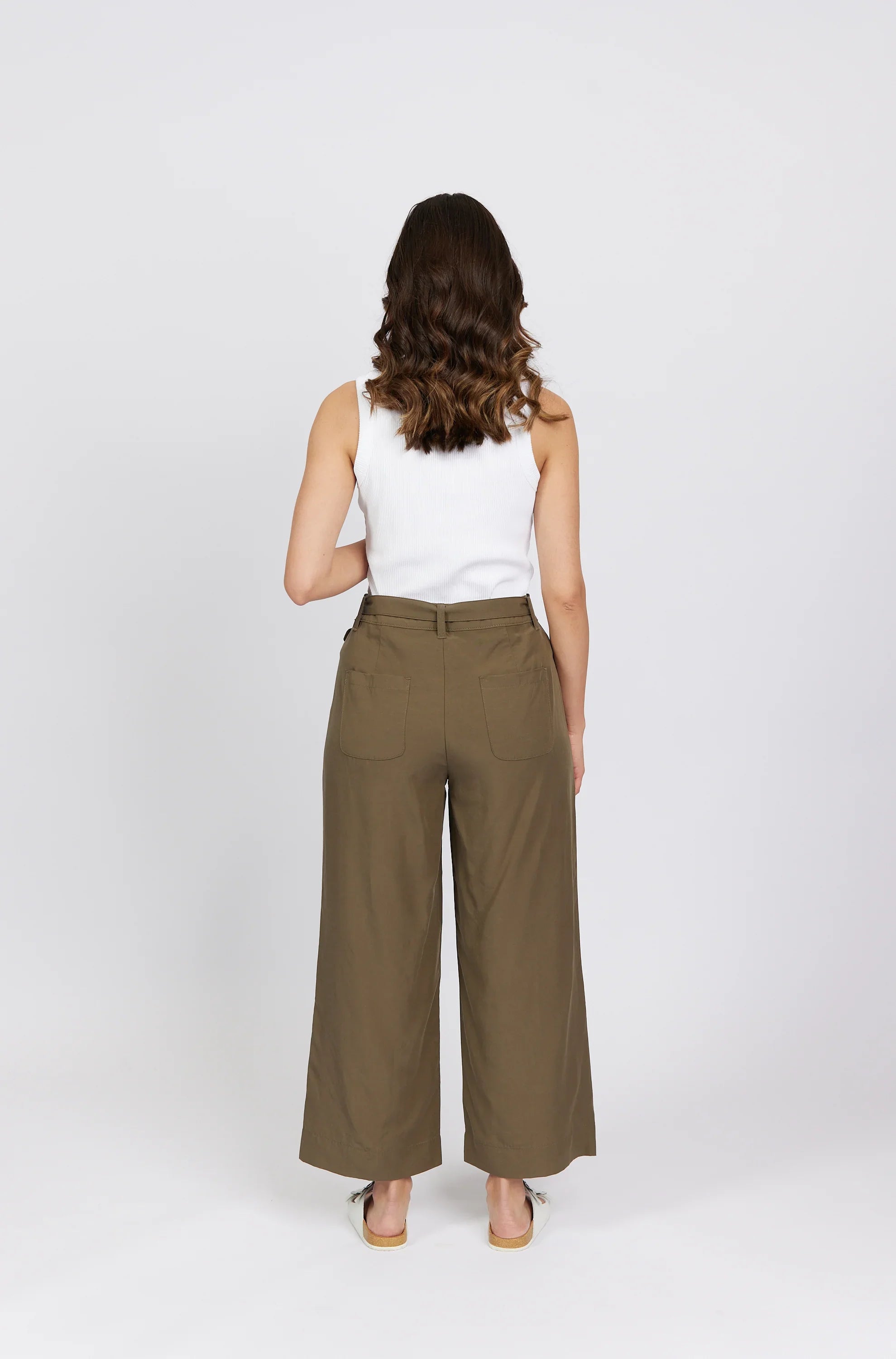 Woman in white tank top and wide leg khaki pants, back view, standing against white background