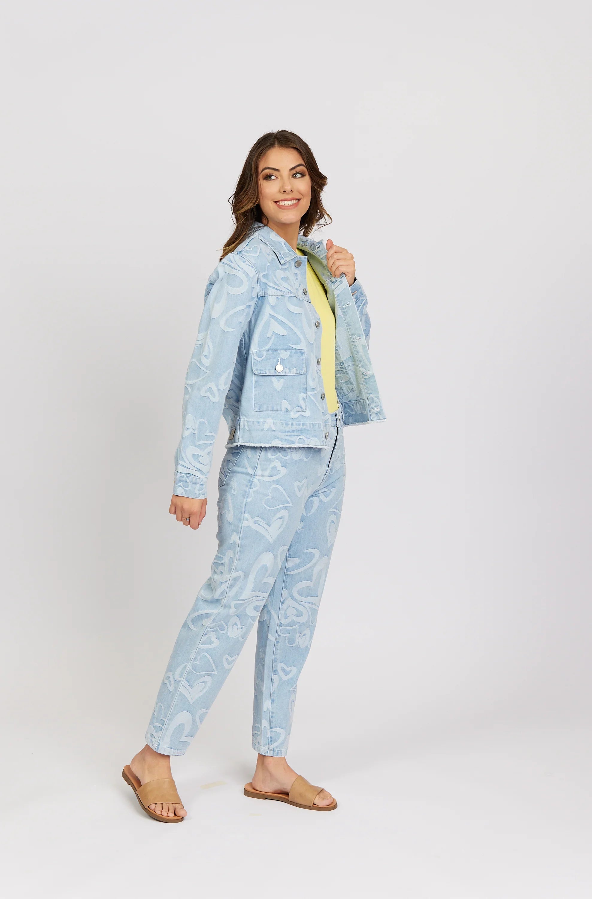 Woman wearing light blue heart-print denim jacket and jeans with tan sandals, studio background