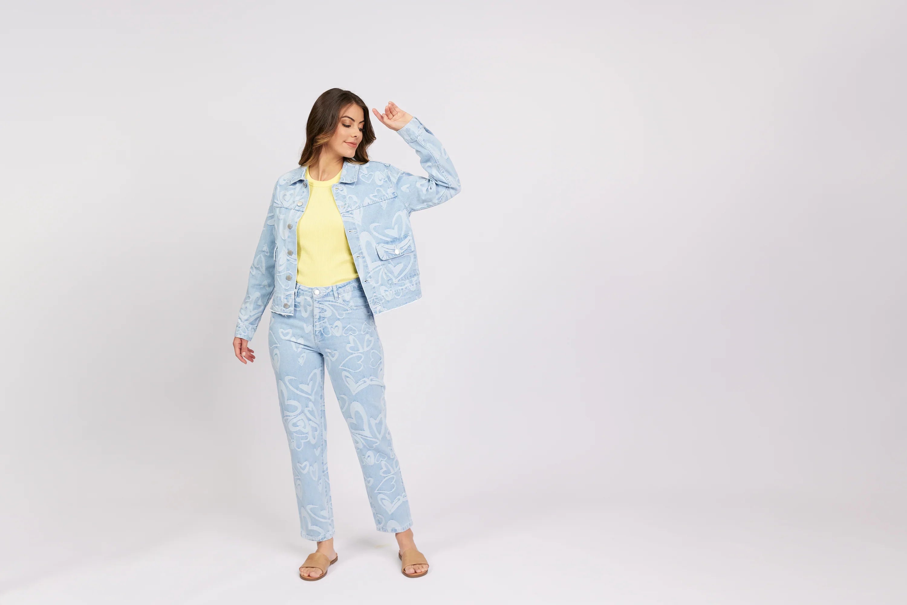 Woman wearing light blue heart print denim jacket and jeans, yellow top, sandals, studio setting