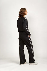 Woman wearing black wide leg pants and track jacket with white side stripes, side view