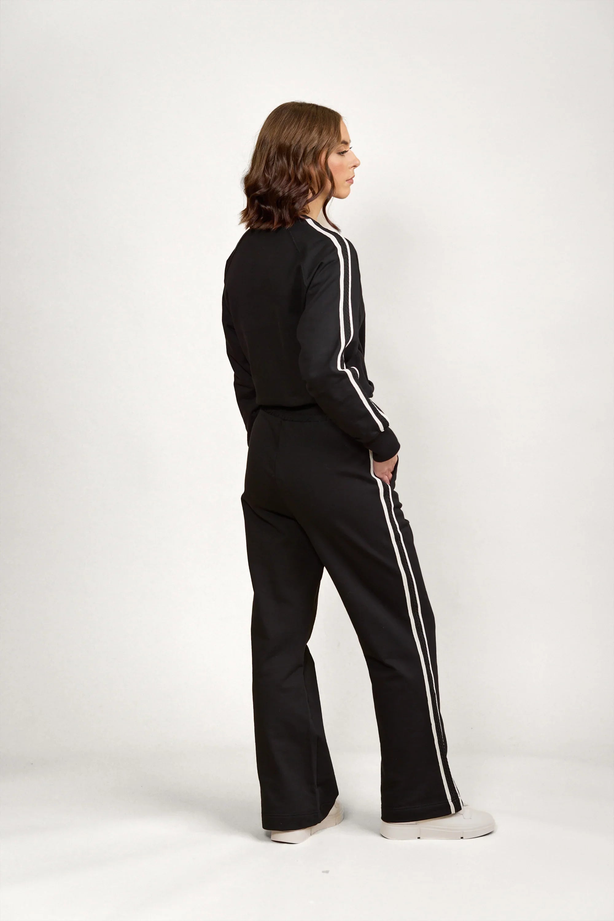 Woman wearing black wide leg pants and track jacket with white side stripes, side view