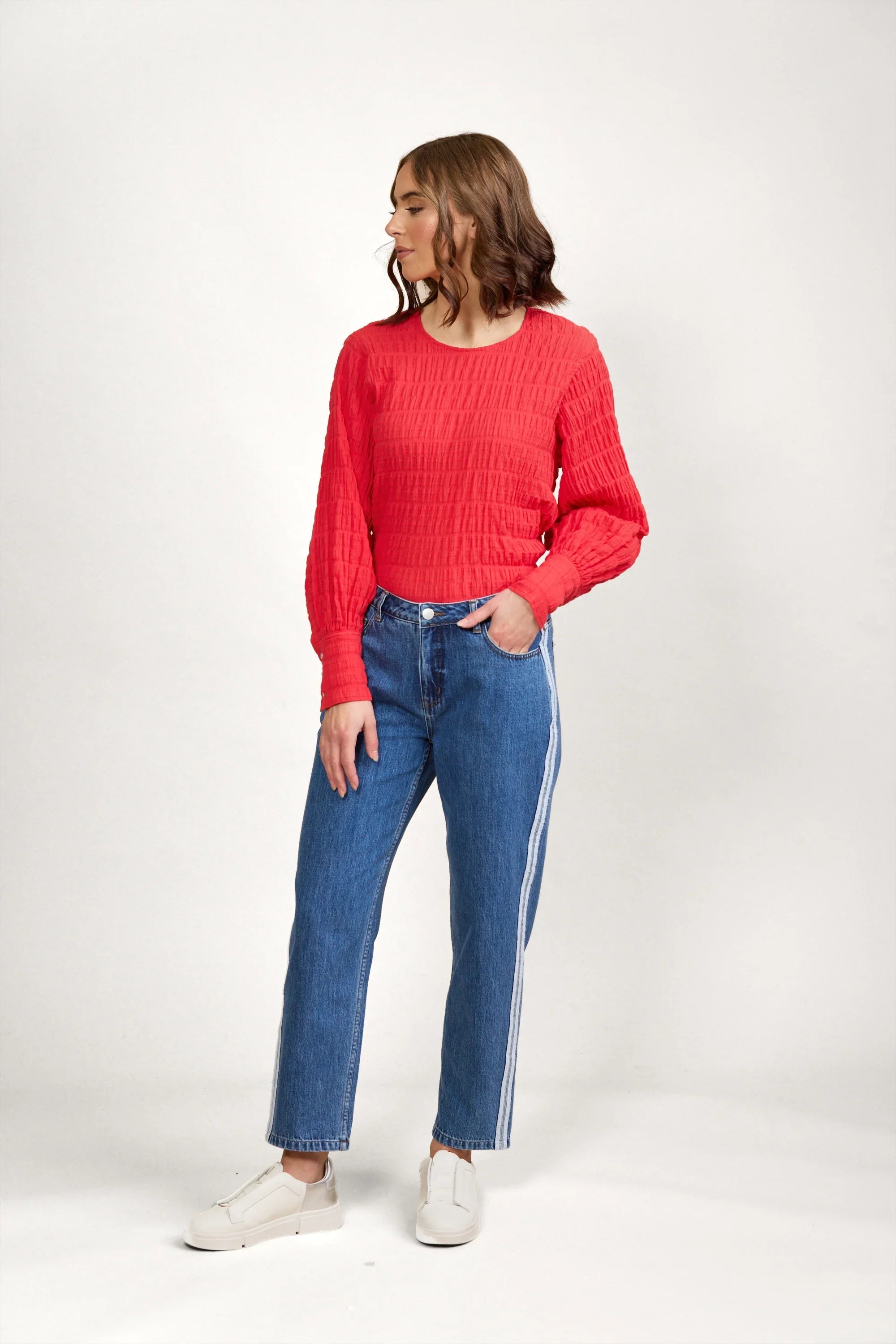 Woman wearing blue straight-leg jeans with side stripe, red textured long sleeve top, white sneakers