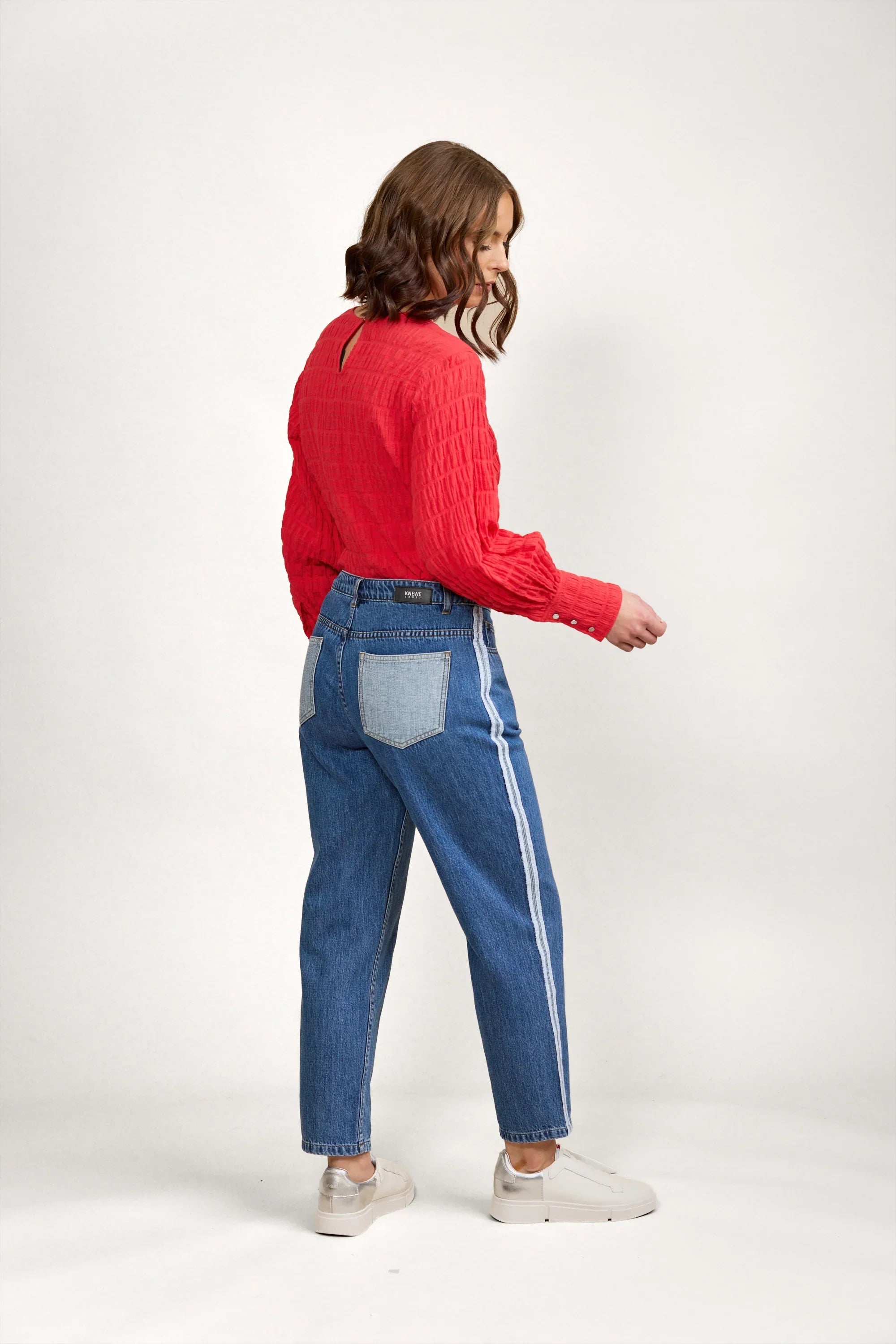 Woman in blue denim jeans with white side stripe and red textured blouse, side view