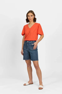 Woman wearing casual V-neck orange top and mid-wash denim Bermuda shorts, studio setting