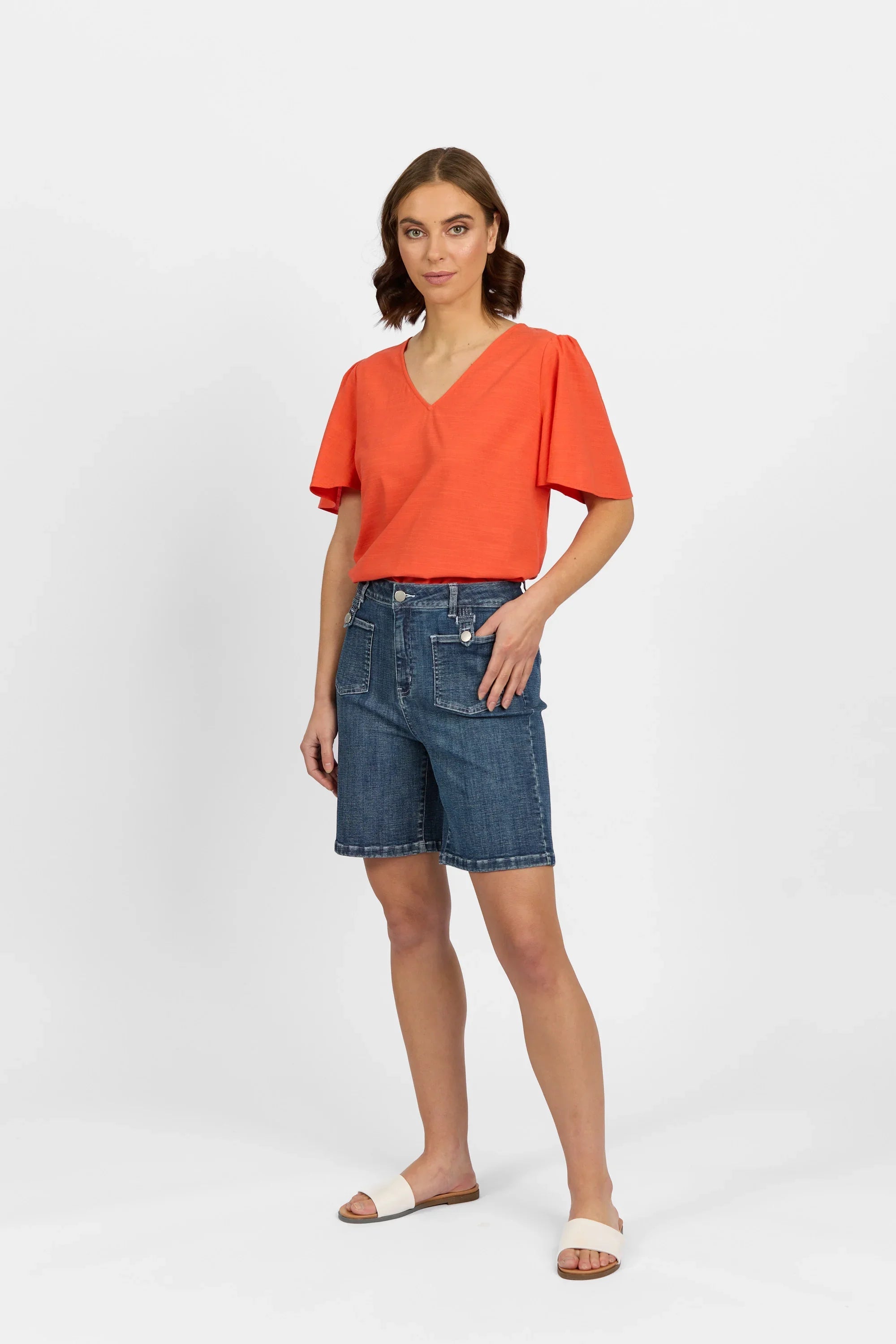 Woman wearing casual V-neck orange top and mid-wash denim Bermuda shorts, studio setting