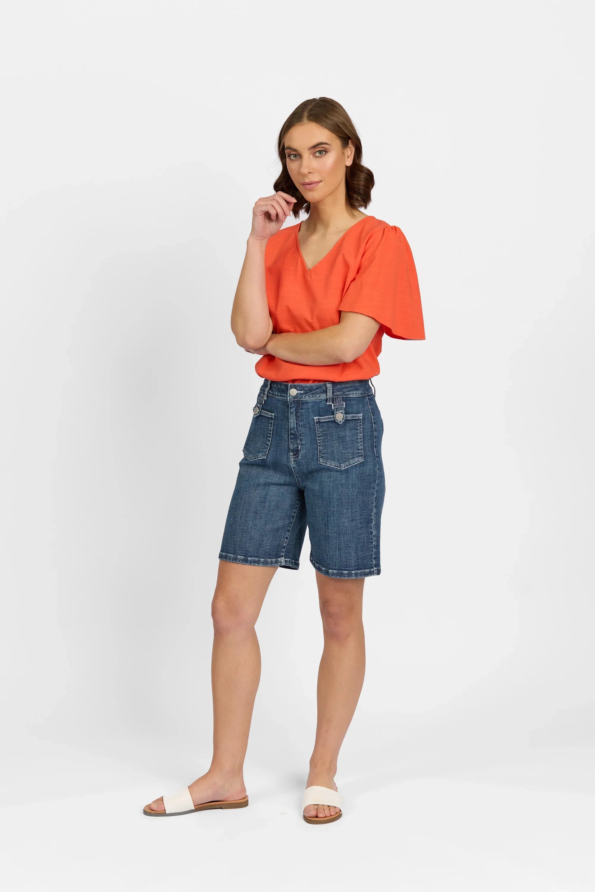 Woman wearing mid-wash denim shorts, orange V-neck top, and white slide sandals