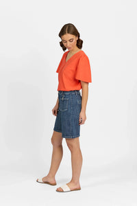 Woman in denim mid-length shorts and orange V-neck short sleeve top, white sandals