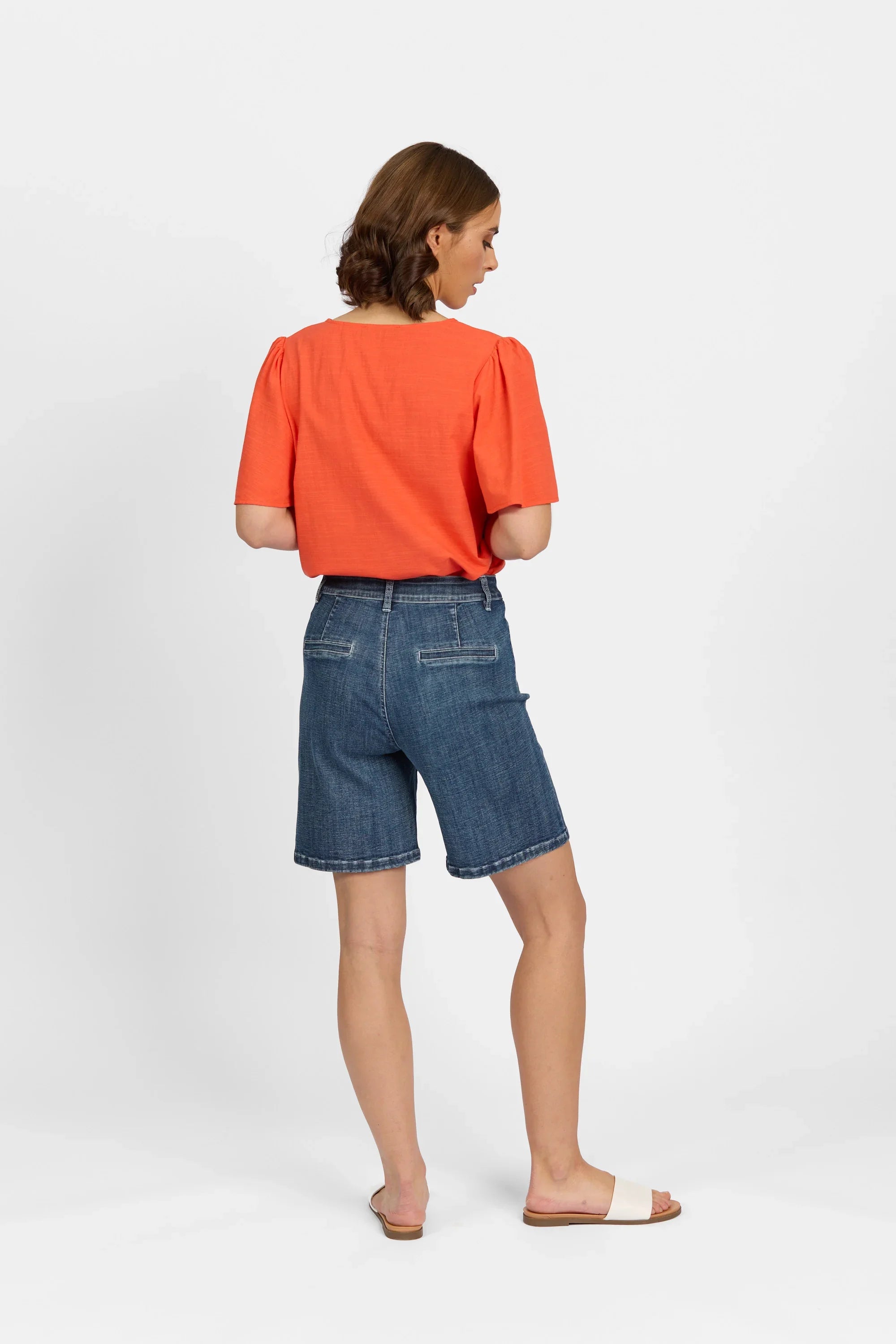Woman wearing mid-wash denim shorts and an orange top, back view, white background
