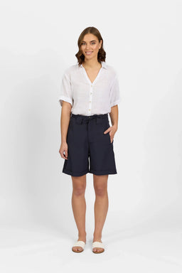 Woman in a white linen shirt and navy knee-length shorts with a tie belt, standing against a plain background.