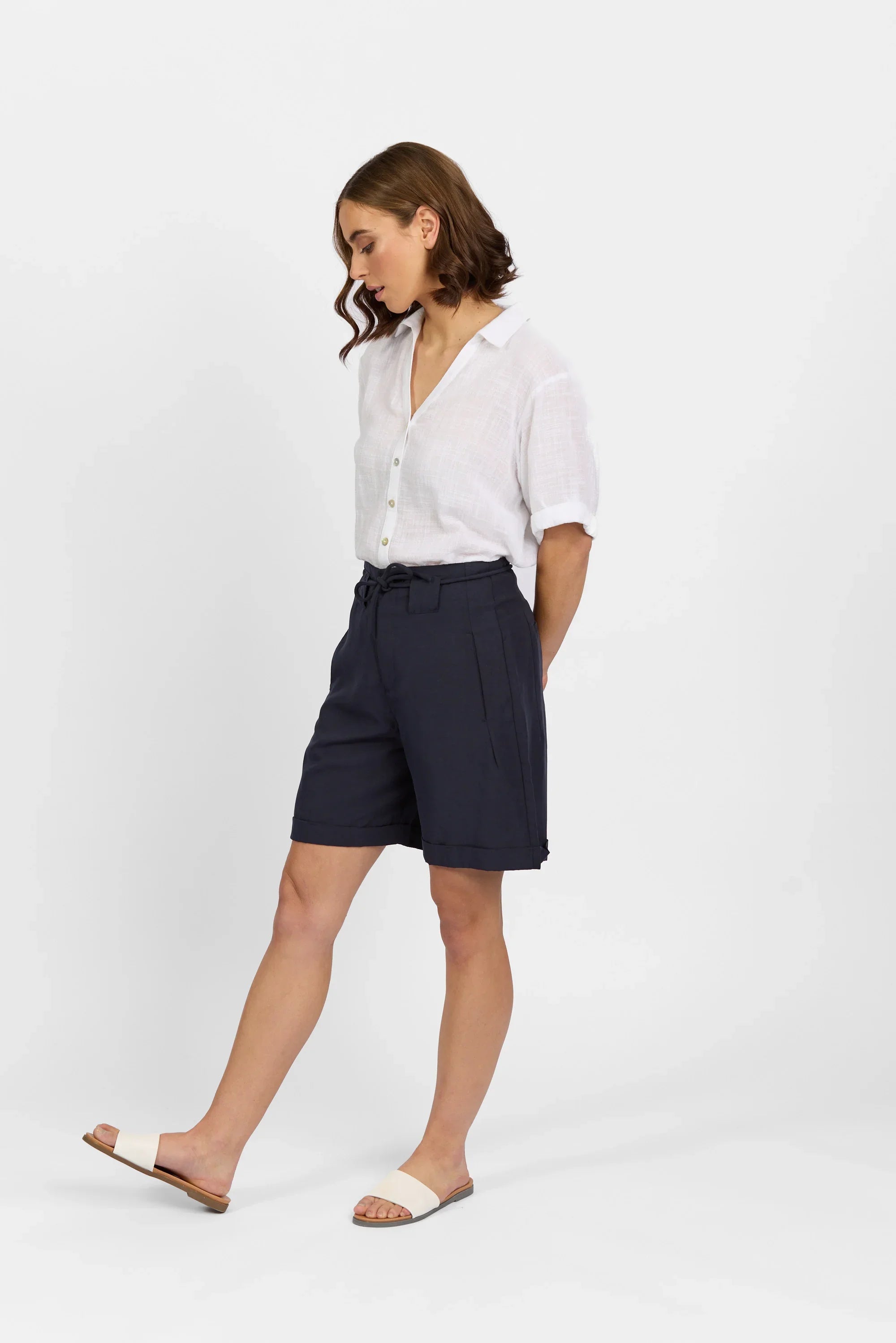 Woman wearing navy tailored shorts, white button-up shirt, and white slide sandals.