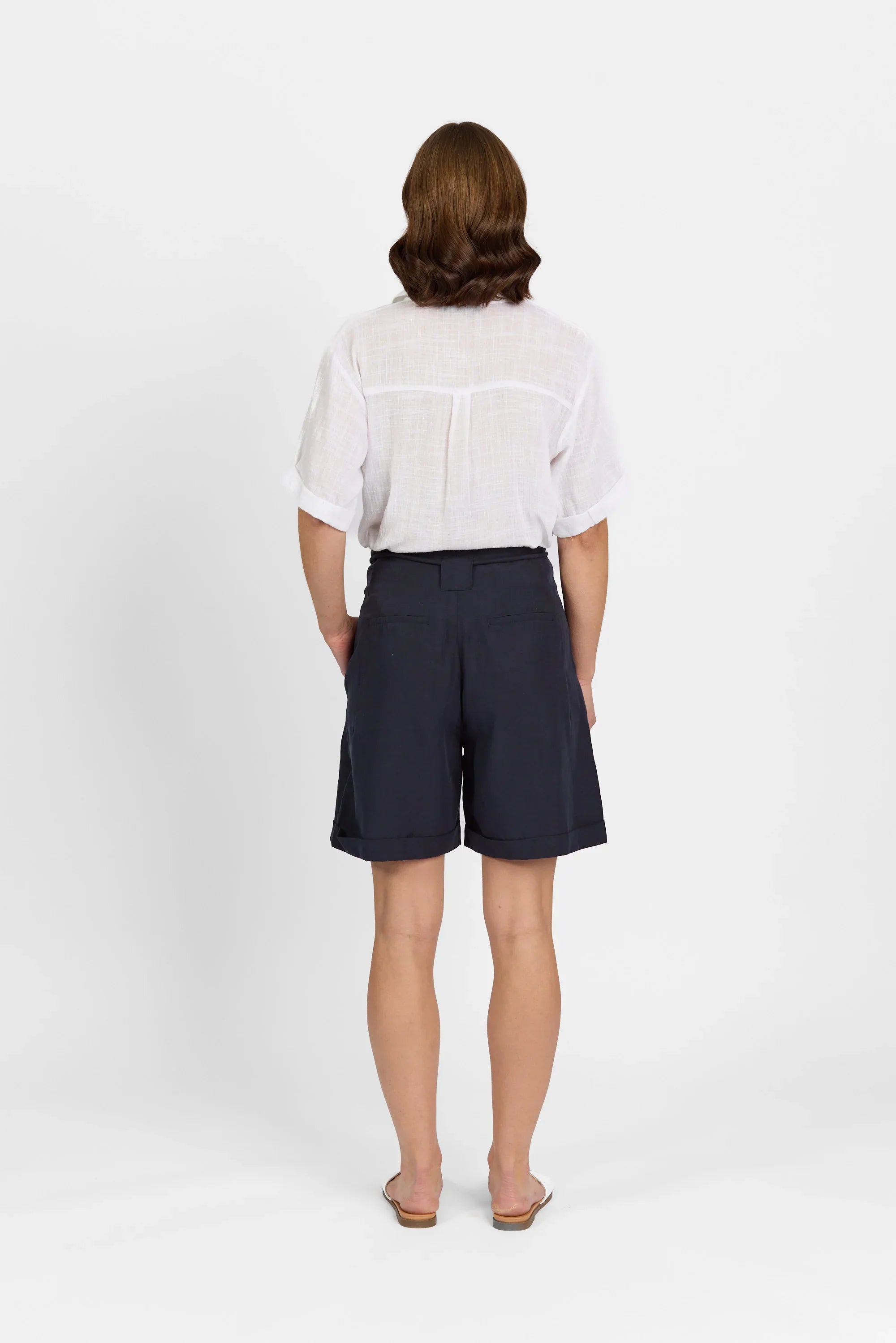 Woman in white short-sleeve shirt and navy tailored shorts, back view, standing indoor