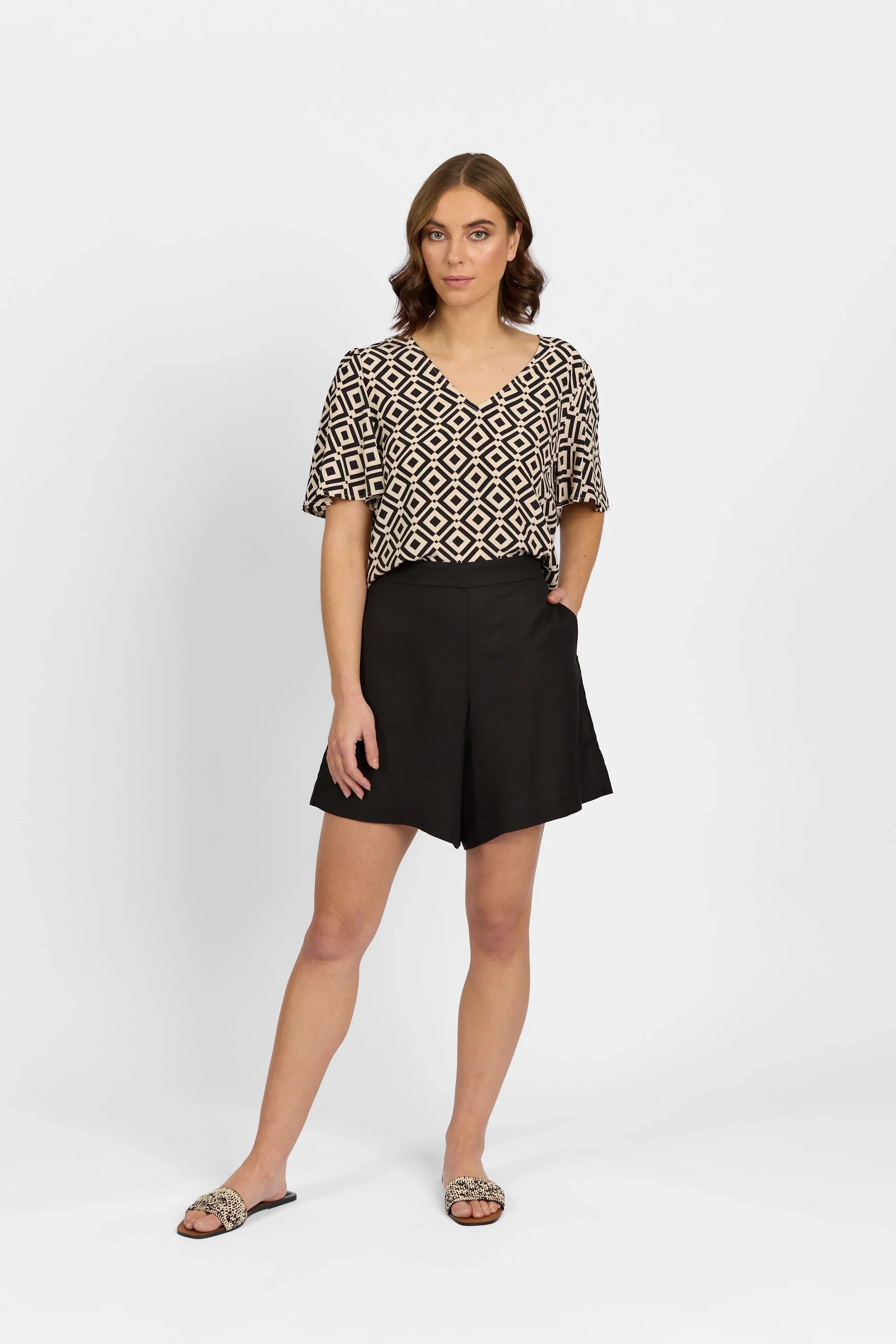 Woman in geometric patterned V-neck top and black shorts, standing against a white background
