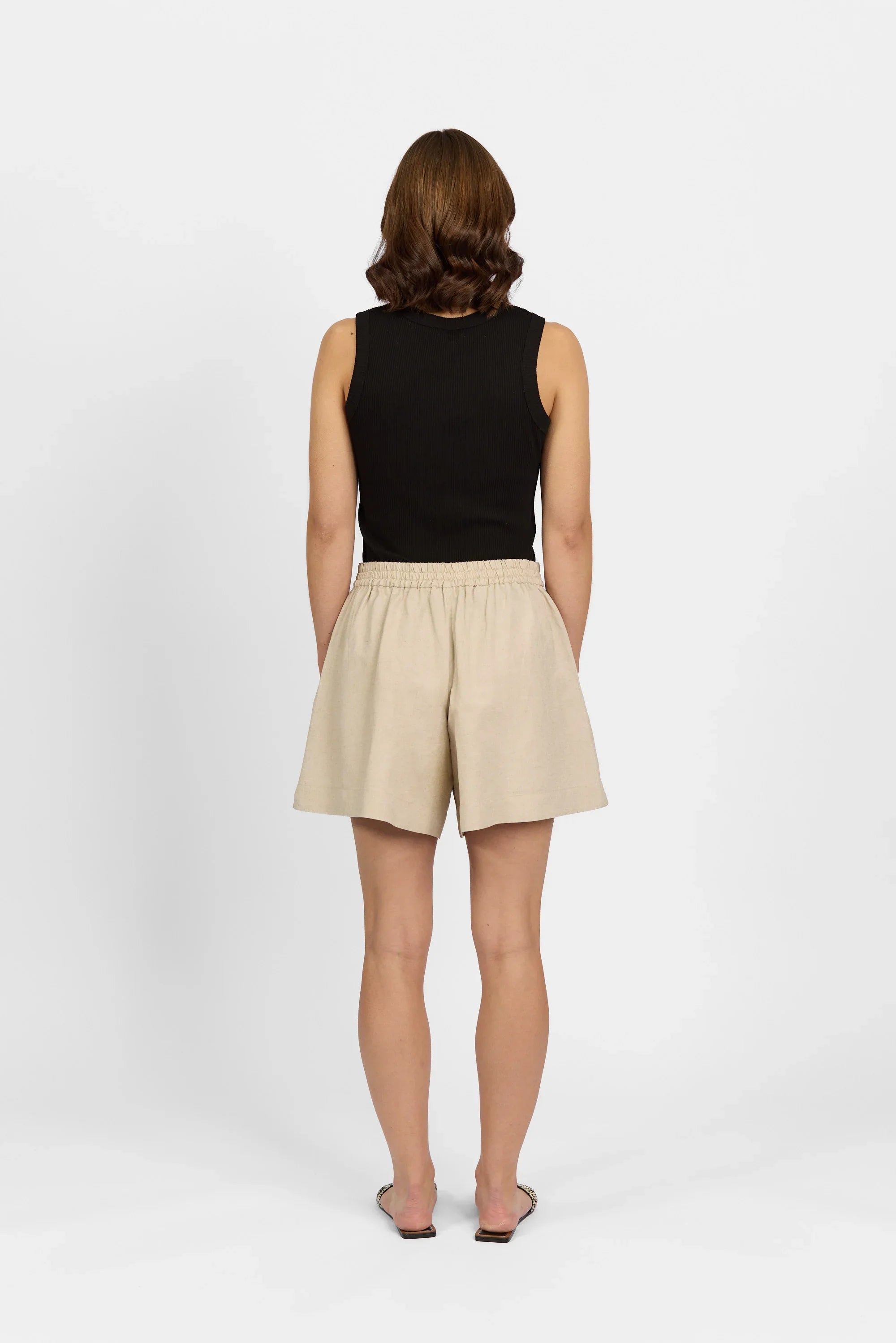 Woman modeling ecru linen shorts with elastic waistband, black sleeveless top, back view