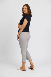 A woman with shoulder-length brown hair stands sideways, wearing a black short-sleeve top and Vassalli Paris Crop Pant in Silver, crafted from stretch fabric, paired with white slide sandals against a plain white background.