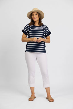 A woman wearing a straw hat, navy and white striped short-sleeve shirt, Vassalli Paris Crop Pant in White, and sandals stands against a plain white background.
