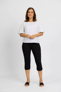 A woman stands against a white background in a loose white short-sleeve top and Vassalli Paris 3/4 Pant - Black, styled with black slide sandals. She smiles slightly, hands lightly clasped in front.