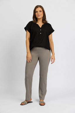 A woman with shoulder-length brown hair and a neutral expression stands against a white background, wearing a black short-sleeve blouse, Vassalli Paris Full Length Pant in Latte, and leopard-print sandals.