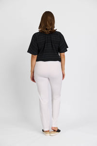 A person with shoulder-length brown hair stands facing away, wearing a black short-sleeve top, Vassalli Paris Full Length Pant in white, and black slip-on shoes against a plain white background.