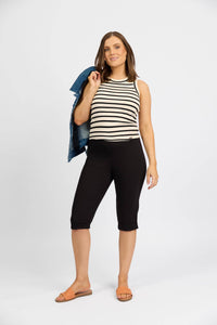 A woman poses against a white background, wearing Vassalli's Paris Short in black, a sleeveless black and white striped stretch top, tan sandals, and holds a blue denim jacket over her shoulder.
