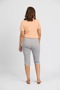 A woman with shoulder-length brown hair stands facing away against a plain white background, wearing a peach short-sleeve shirt, sandals, and Vassalli’s Paris Short - Silver mid-rise stretch capri pants.