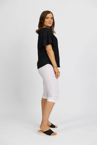 A woman poses sideways against a light background, wearing Vassalli's Paris Short in white, a black stretch short-sleeve top, and black platform sandals.