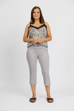 A woman stands against a white background, wearing a black-and-white patterned sleeveless top with lace trim and Vassalli Paris 3/4 Cargo Shorts in Silver. She smiles warmly, hands clasped, finishing the look with black slide sandals.